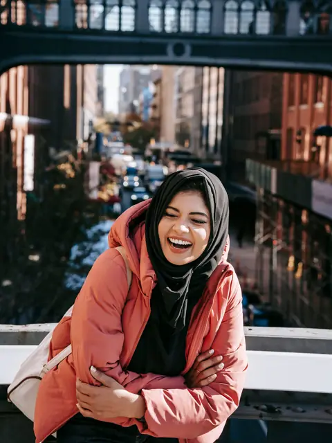A woman in a headscarf and a red jacket laughing on a bridge above a busy city street